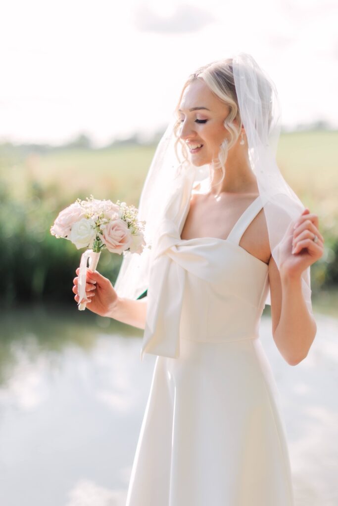 Blonde bride looking down at her bouquet by a lake, soft bridal hair and makeup in Leicester